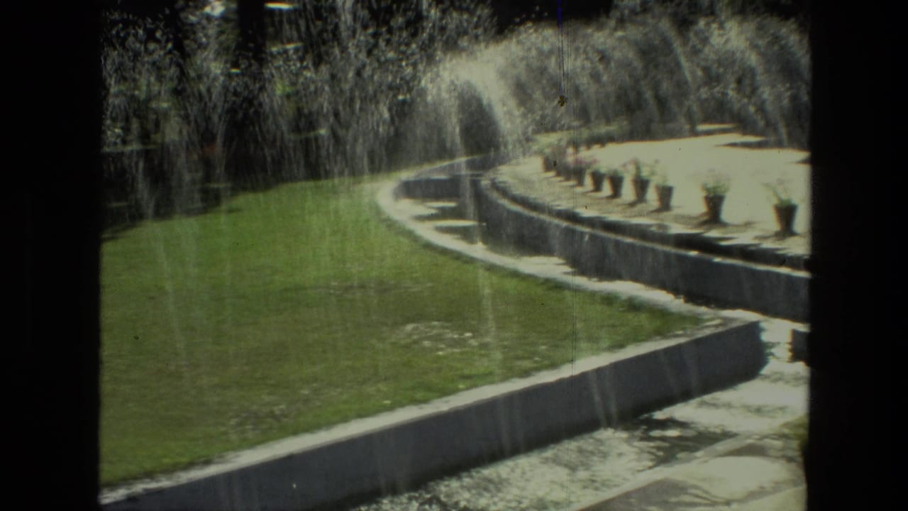 Sunlit India garden scene on 8mm film, with fountain spray crossing a lawn and a raised curved terrace lined with planters.