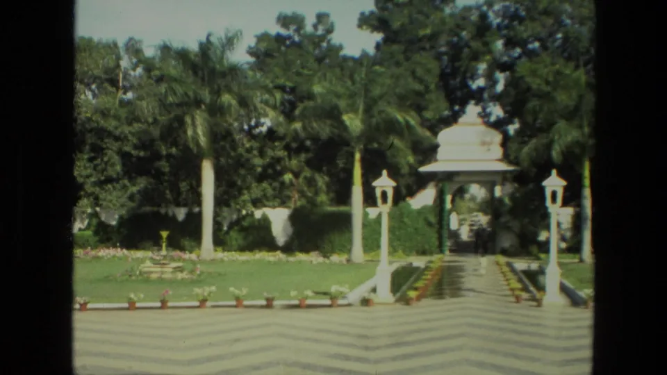 1974 India home-movie frame of a manicured garden with palms, lawn, white lamp posts, potted flowers, and a pavilion entrance.