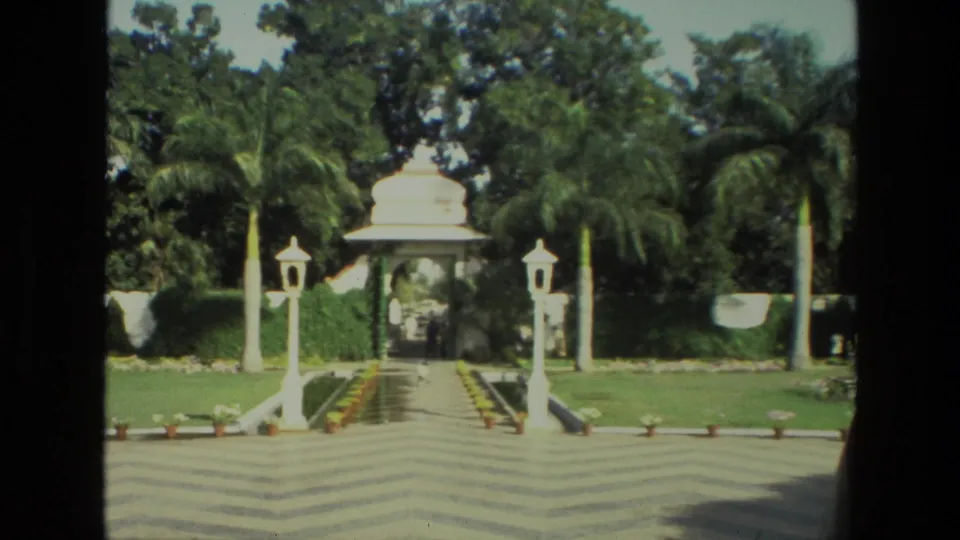 A formal garden walkway in India, filmed in 1974, shows white lamp posts, palms, hedges, and a pavilion at the far end.