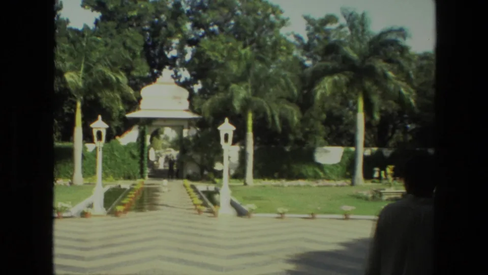 1974 India 8mm frame showing a chevron-patterned courtyard path, white domed gateway, palms, lamp posts, and a person at the right edge.