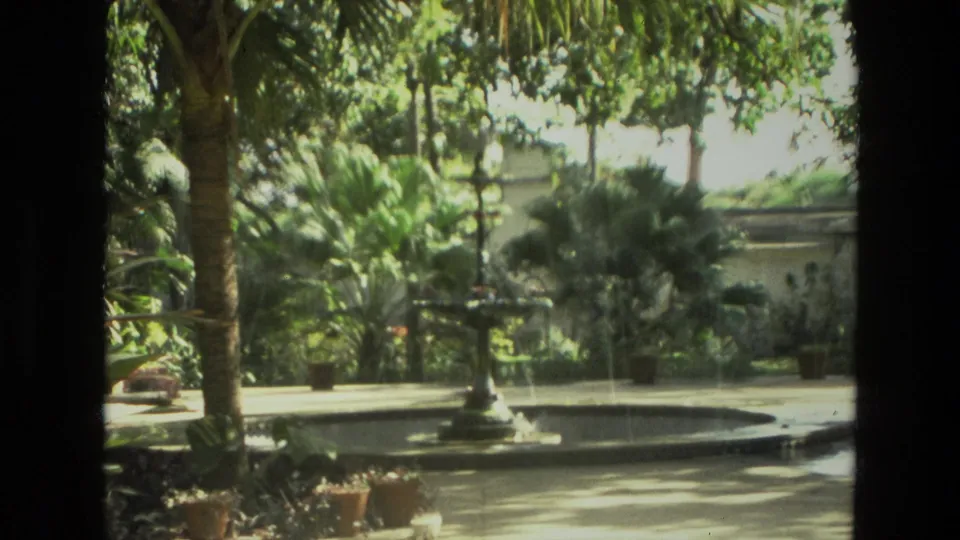 1974 India 8mm frame showing a fountain basin in a landscaped courtyard, with palms, potted plants, and dark framing at the sides.