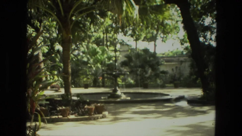 1974 India home-movie frame of a garden fountain, circular pool, palms, potted plants, and bright courtyard light beyond a shaded edge.