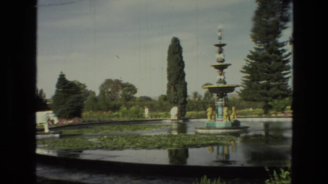 1974 India 8mm frame of an ornate multi-tier fountain reflected near a lily-pad pond, with tall trees in the garden.