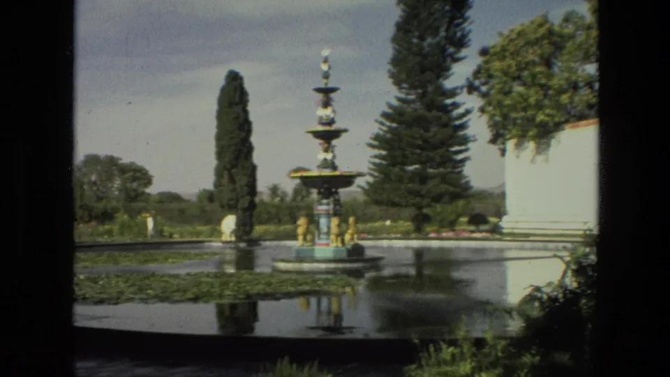1974 India 8mm view of the fountain across a pond with lily pads, trees, and part of a pale garden structure at right.