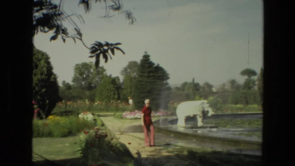 1974 India 8mm frame with an adult woman in red near a pond, white elephant sculpture, flower beds, trees, and garden path.