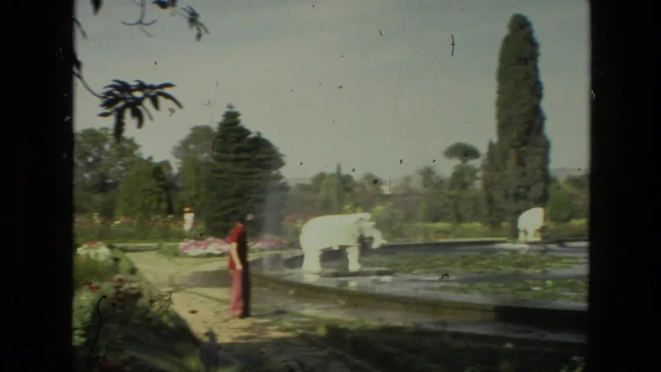 In 1974 India home-movie footage, a visitor in red pauses beside a pond with a white animal sculpture, tall trees, paths, and manicured planting around the water.