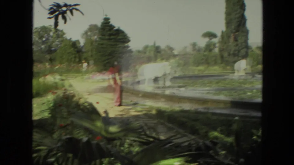 A 1974 India garden scene shows a visitor near a pond with a white sculpture and water spray, framed by tall trees, shrubs, and formal landscaping.