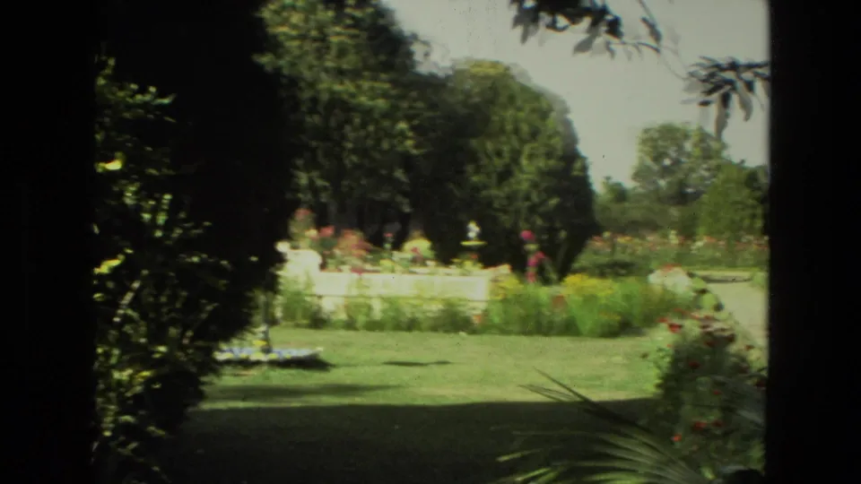 A wider 1974 India home-movie view looks across lawn and planted beds toward visitors walking among trees and ornamental garden features.