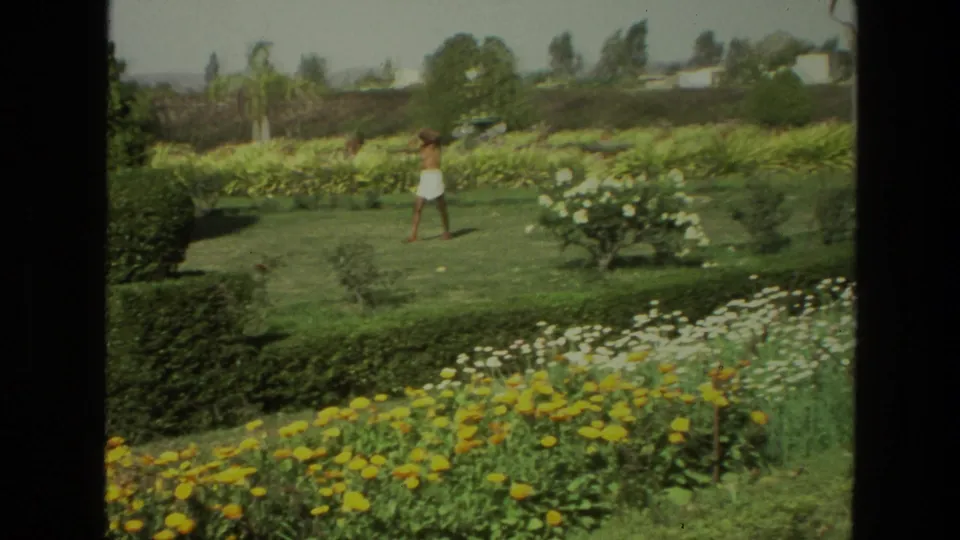 1974 India 8mm frame showing a person crossing a garden lawn bordered by hedges and flower beds under bright daylight.