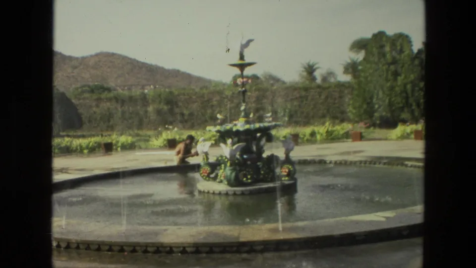 1974 India garden frame with a person seated by a circular ornamental fountain, water jets, trees, and low hills beyond.