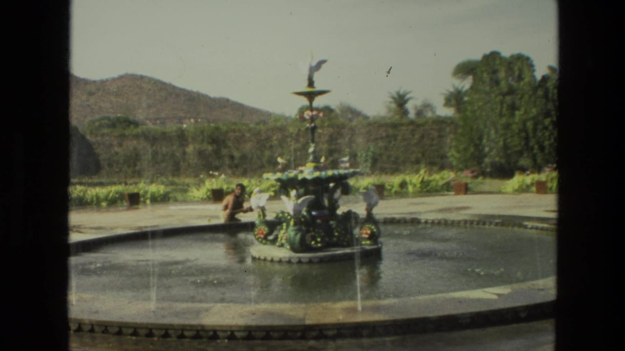 1974 8mm India travel frame showing a decorative fountain in a shallow garden pool with a person nearby and low hills in the background.