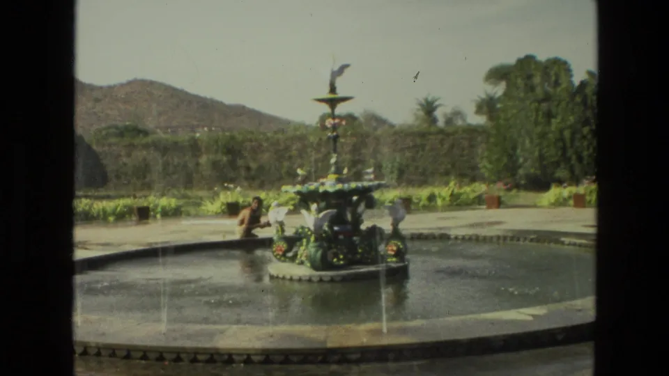 1974 8mm India travel frame showing a decorative fountain in a shallow garden pool with a person nearby and low hills in the background.