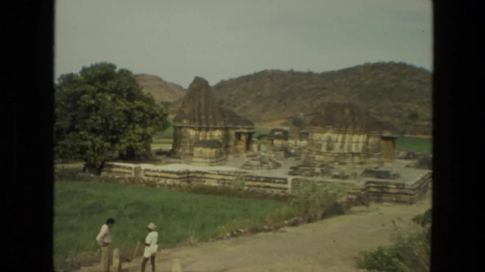 1974 India travel frame with two people walking on a path near stone temple buildings, greenery, a large tree, and low hills.