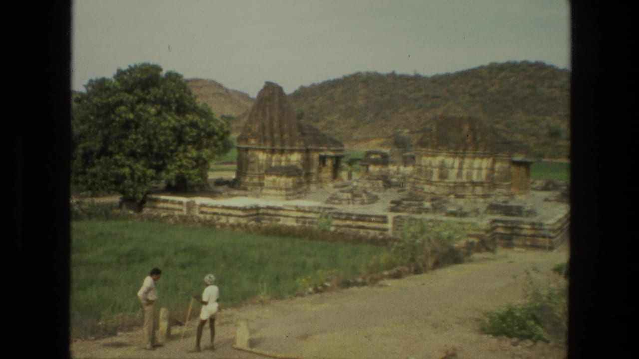 Two men stand on an unpaved path near stone temple ruins and a large tree.