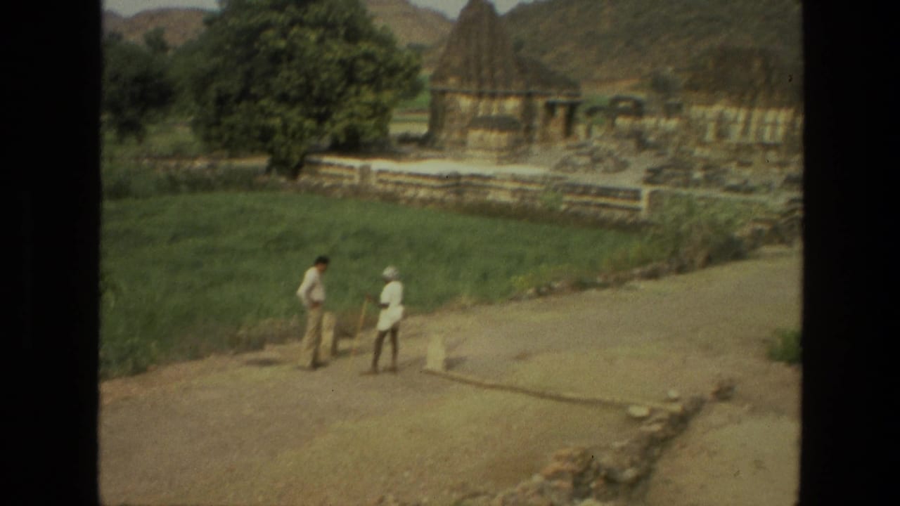 1974 India 8mm frame showing two men on a rural path below stone temple ruins, with grass and low hills in the background.