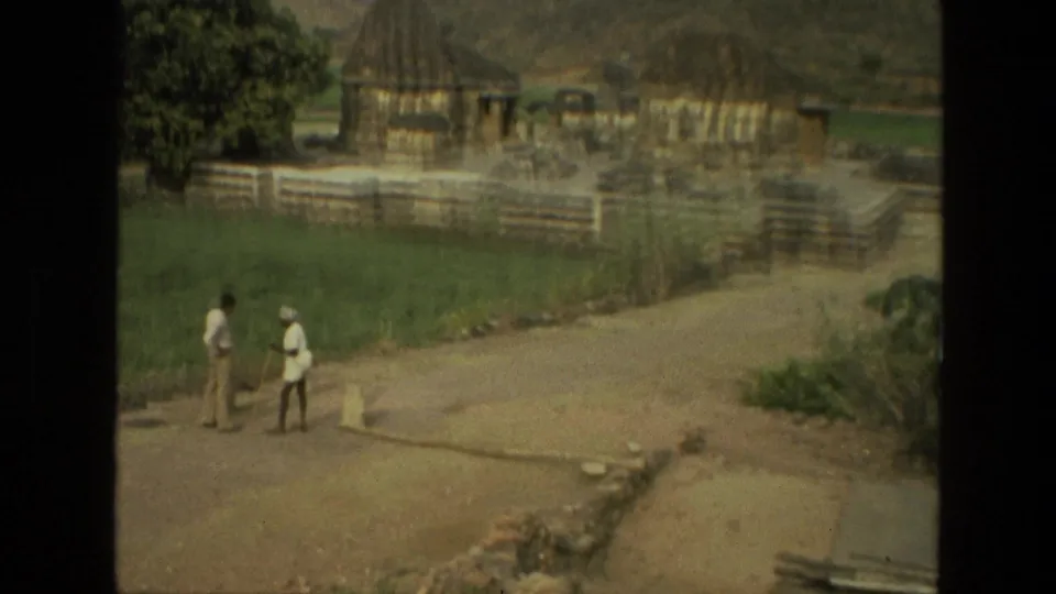 Soft 8mm frame from India in 1974 showing two men near grass and temple ruins, with handheld blur and faded color.