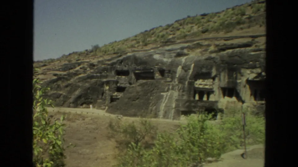 1974 India travel frame showing hillside rock-cut cave architecture with carved openings, stone steps, and sparse vegetation.