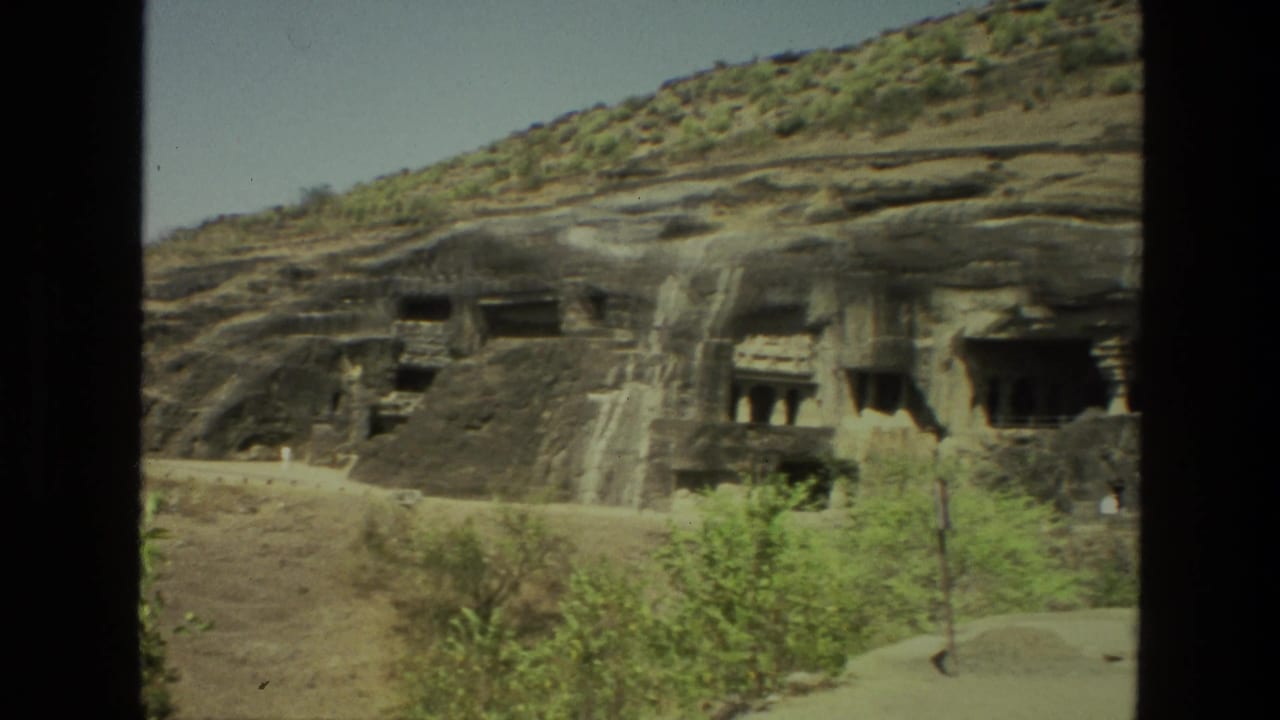 Rock-cut cave temple facades in a dry hillside with sparse shrubs and a sunlit path.