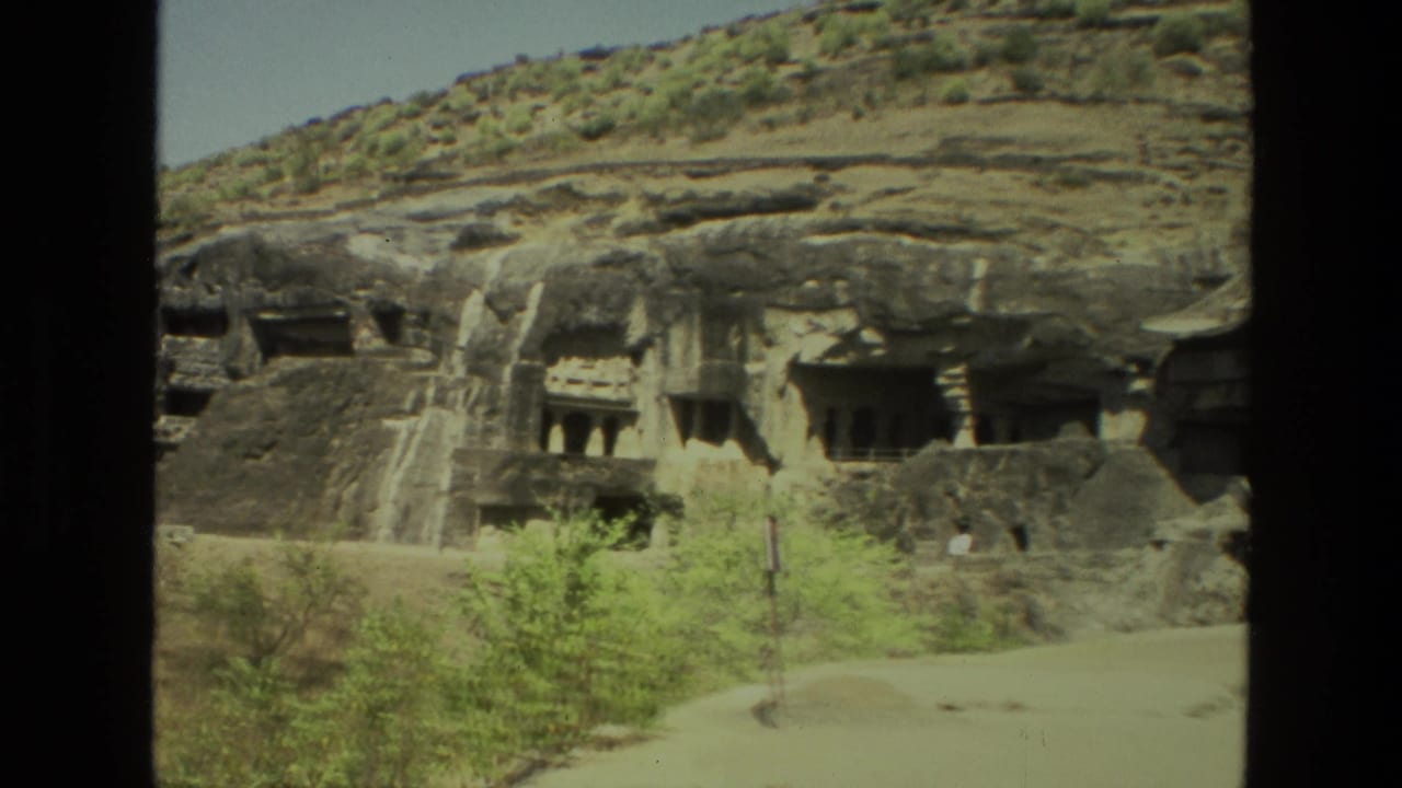 Carved cave entrances and rocky ledges above a pale path with shrubs in front.