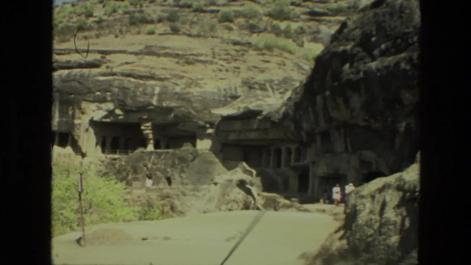 1974 India home-movie frame showing a wider cave temple view with dry walkway, shrubs, carved openings, and small visitors.