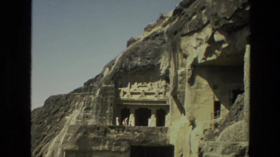 Closer 1974 8mm view of a carved cave temple facade in India, with dark openings and small visitors at the entrance.