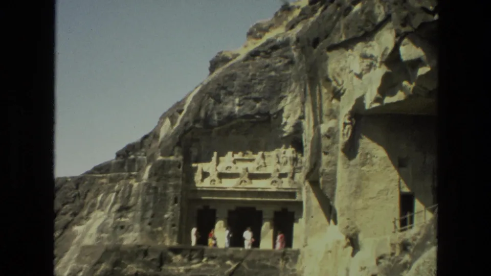1974 India 8mm frame of visitors on a stone ledge before an ornate rock-cut cave or temple entrance.