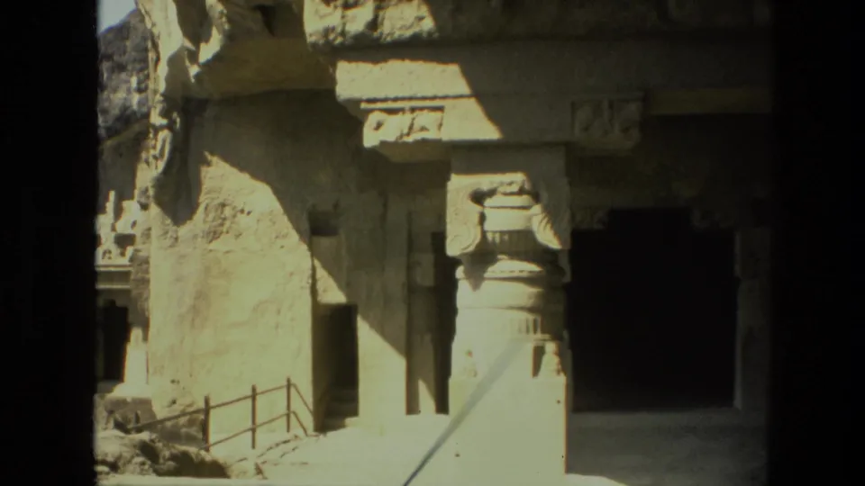 1974 India travel footage frame of carved rock-cut stonework, a railing, and a dark cave entrance in strong sunlight.