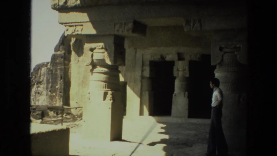 1974 India home-movie frame showing a visitor beside carved rock-cut pillars and shadowed cave openings.