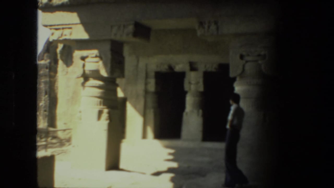 Sunlit stone temple interior with pillars, dark openings, and a lone visitor in shadow.