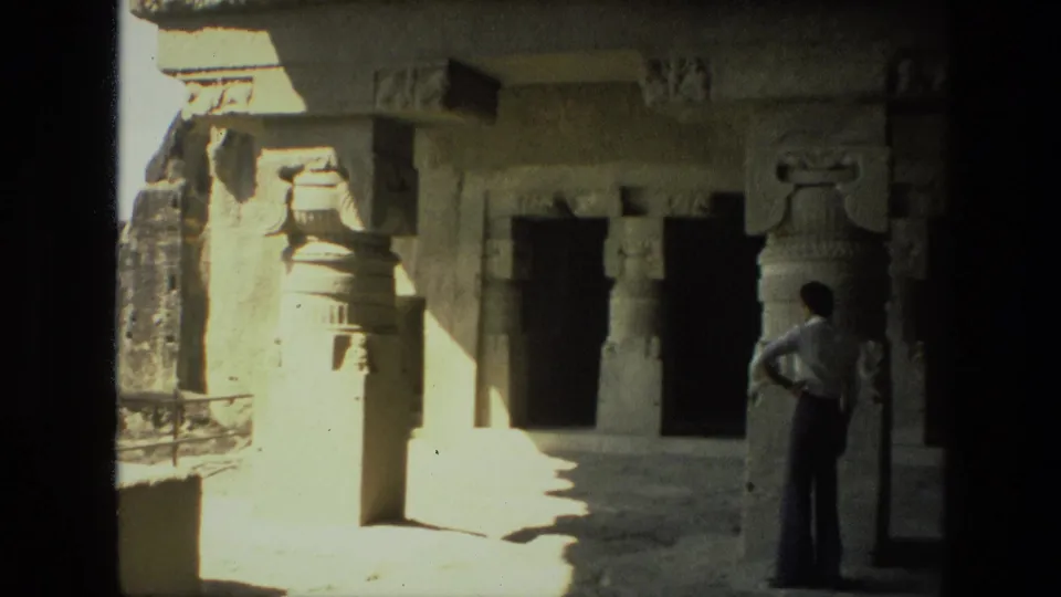 1974 India home-movie frame of a visitor looking across a stone ruin interior with carved pillars and deep shadowed entrances.