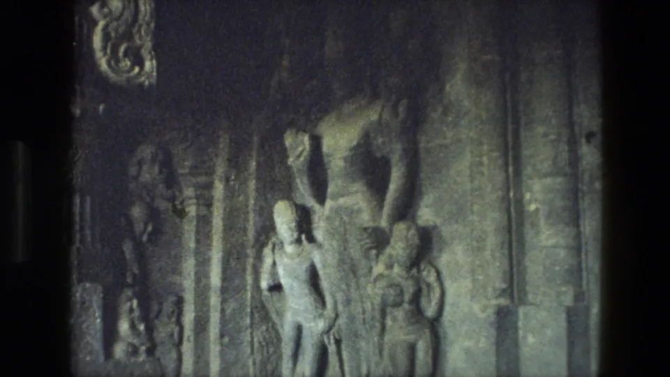 1974 India home-movie frame of a carved stone wall with human-like figures, relief details, and muted low-light film texture.