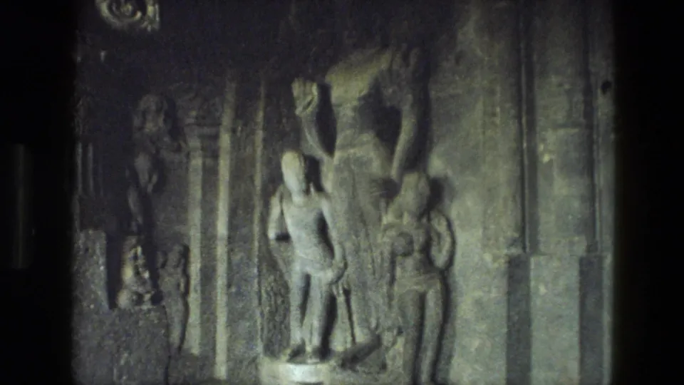1974 8mm frame from India showing carved human figures in stone relief within a dim temple interior.