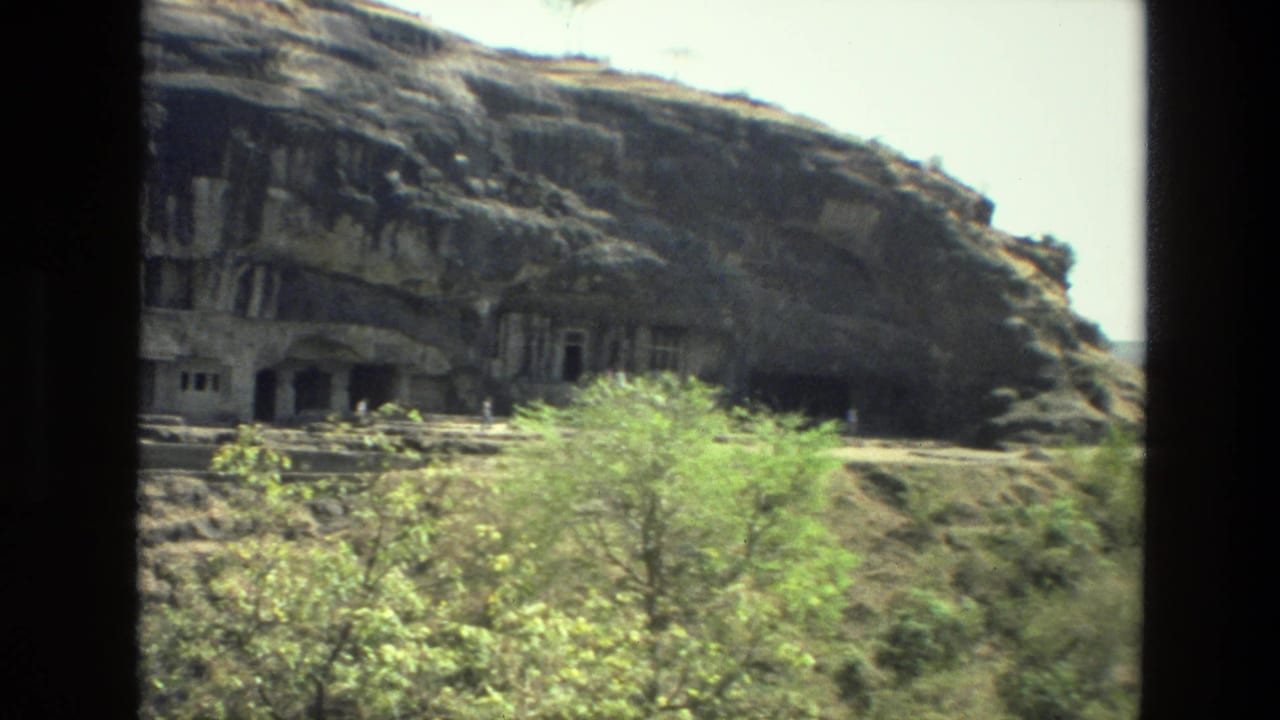 1974 8mm India travel frame showing rock-cut cave entrances across a rugged hillside with sparse green vegetation below.
