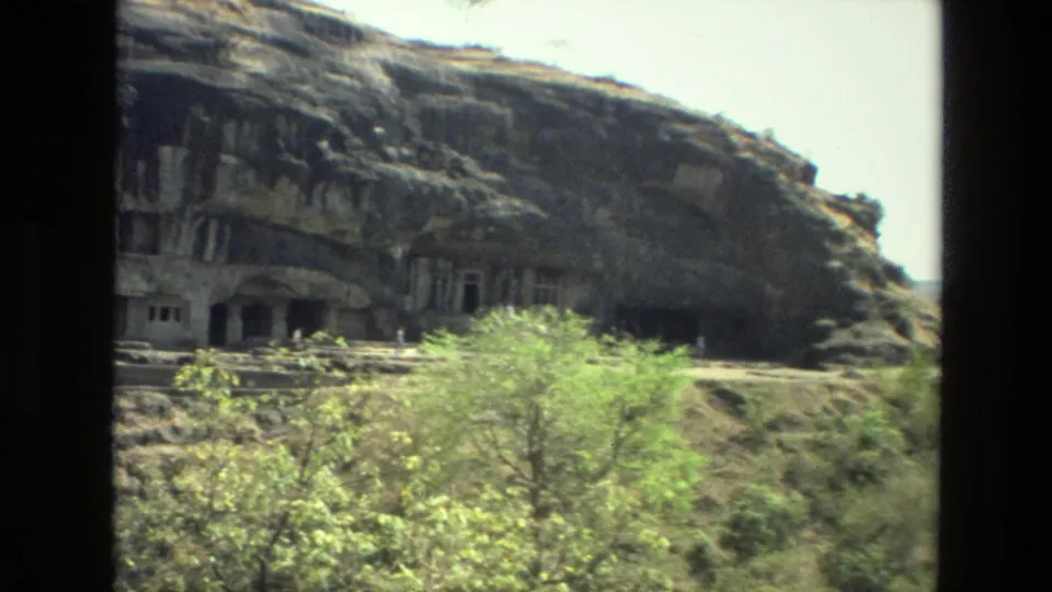 1974 8mm India travel frame showing rock-cut cave entrances across a rugged hillside with sparse green vegetation below.