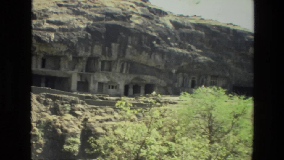 1974 8mm India travel frame showing a closer hillside view of rock-cut openings, ledges, and scrubby greenery in front.