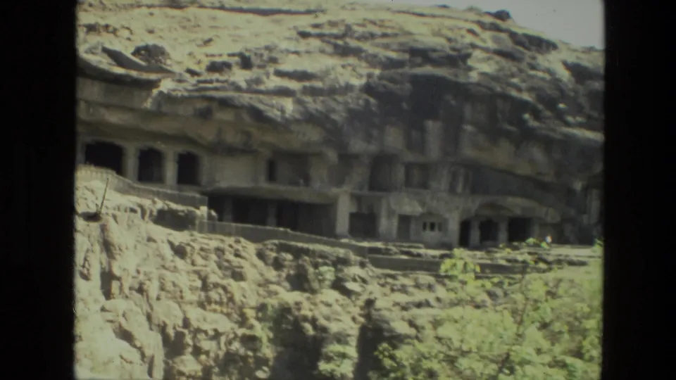 1974 8mm India travel frame showing rock-cut cave doorways beneath an uneven stone overhang, with rough masonry and greenery below.