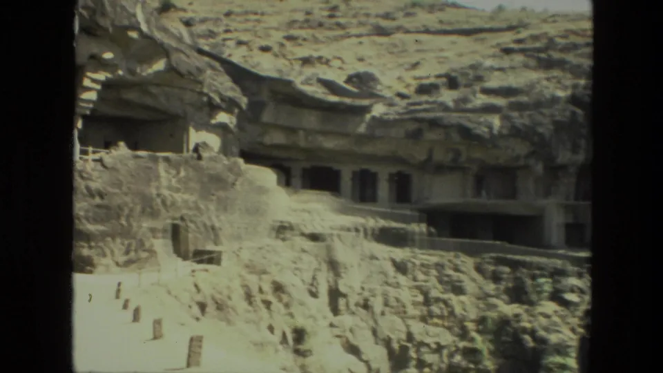 1974 India 8mm view of rock-cut cave temple structures with steps, railings, and shadowed chambers in the cliff.