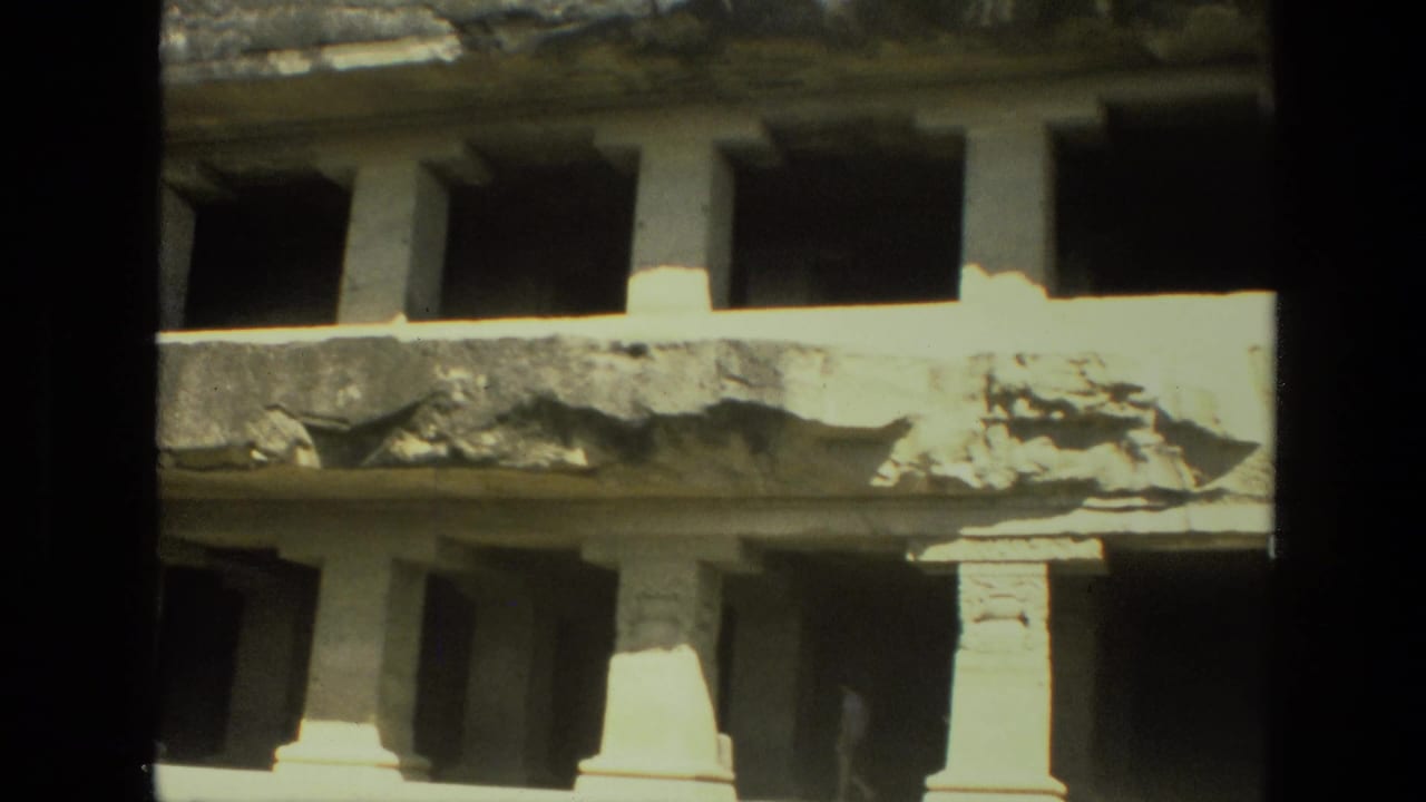 Weathered stone facade with dark openings, columns, and strong sunlight.