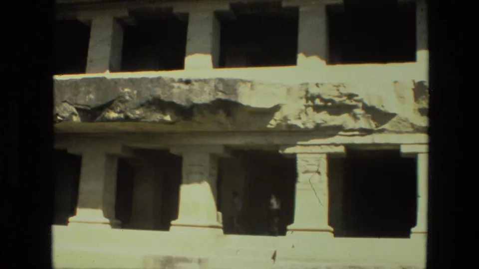 1974 India home-movie frame of a worn stone structure, with column supports below a damaged ledge and dark recessed rooms.