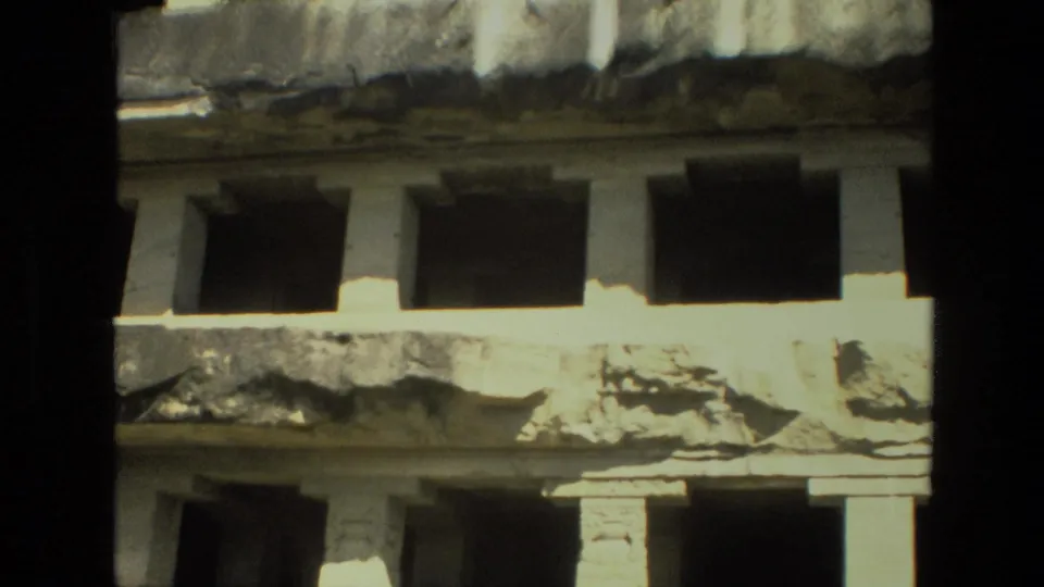 1974 India travel footage frame showing layered weathered stonework, columned supports, and dark openings along the lower level.