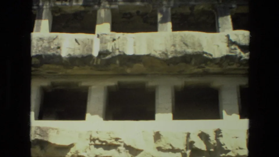 1974 India home-movie frame of a weathered multi-level stone facade, with small visitors silhouetted near the lower archway area.
