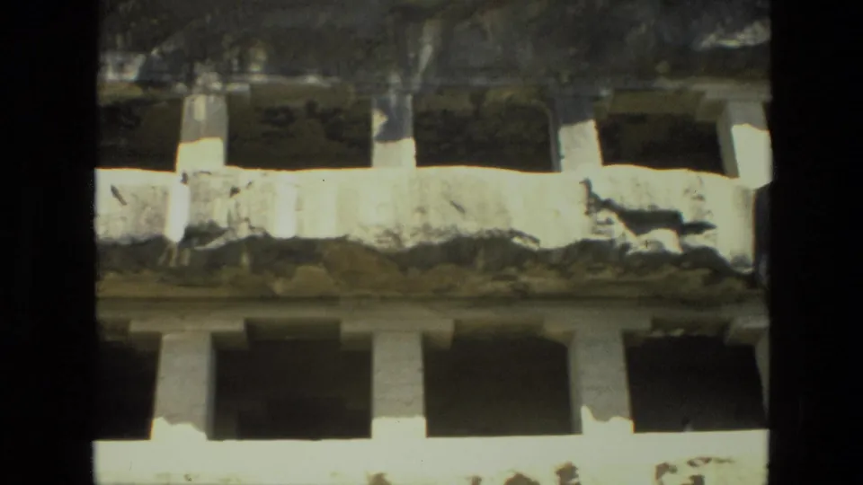 Grainy 1974 8mm view in India showing weathered cave temple stonework with rectangular openings and eroded architectural details.