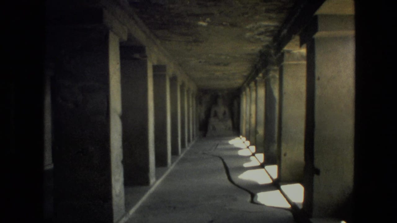 Dim 1974 India 8mm corridor view with stone pillars, side openings, floor shadows, and a distant seated Buddha statue.