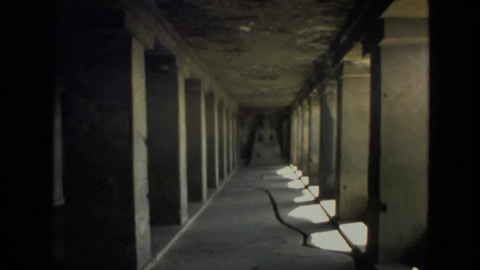 Dim 1974 India 8mm corridor view with stone pillars, side openings, floor shadows, and a distant seated Buddha statue.
