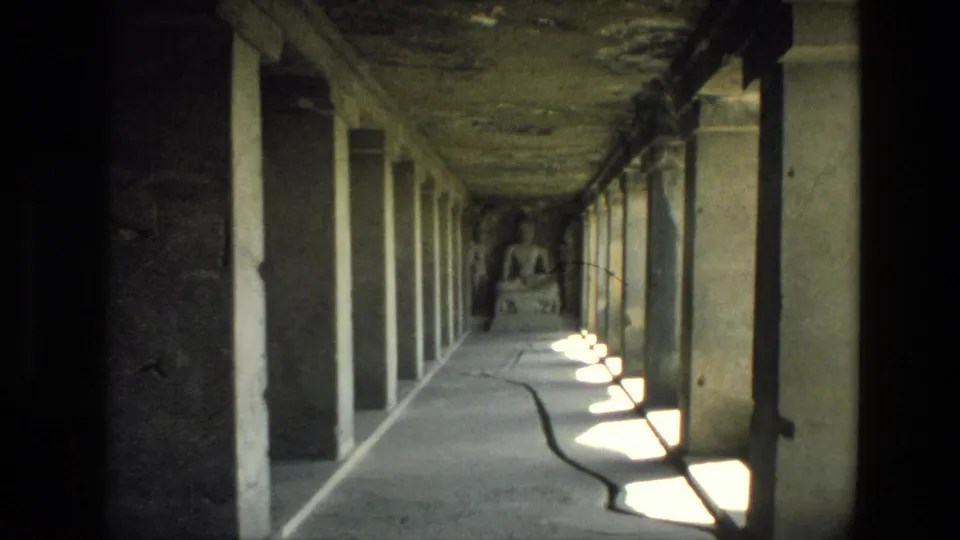 Visitor-shot 8mm frame from India in 1974 showing a cave temple corridor lined with pillars and a seated Buddha statue beyond.
