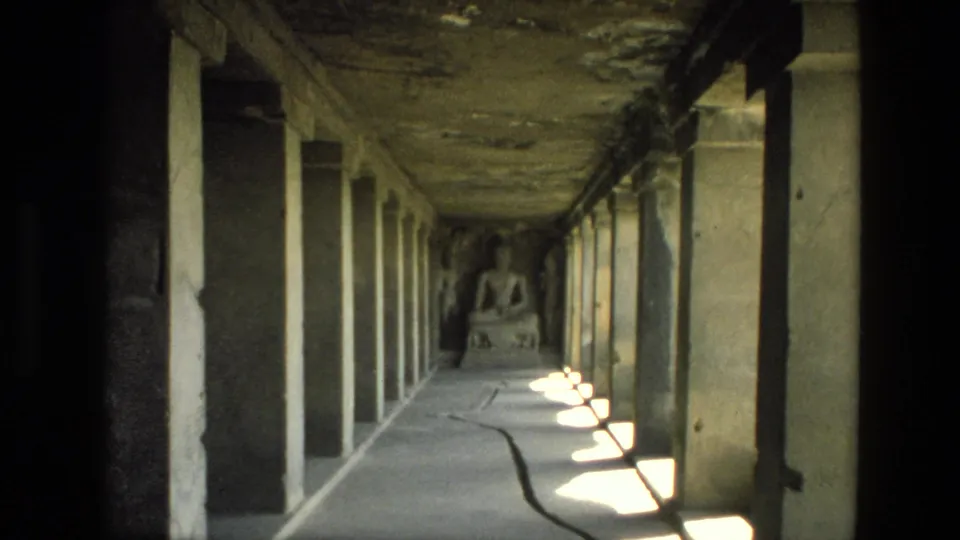 Grainy 1974 8mm frame in India with the corridor depth compressed around pillars, side light, and a seated Buddha statue.