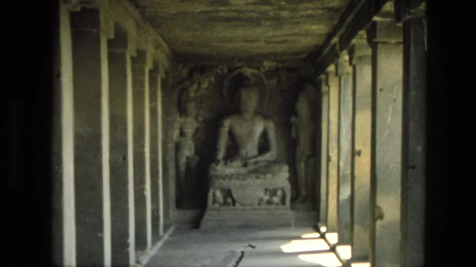 1974 India 8mm frame of a shadowed seated religious statue at the end of a stone temple corridor with pillars and sunlight.