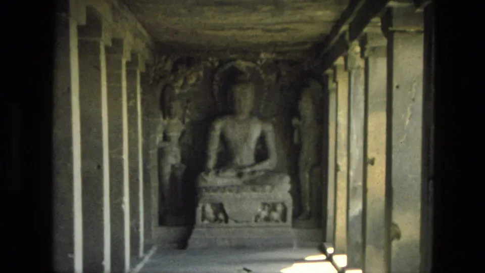 1974 India 8mm frame showing a seated Buddha statue in a rock-cut cave temple corridor with pillars and side carvings.