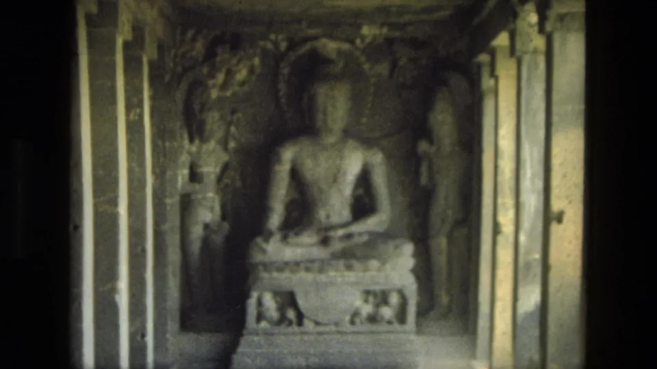 1974 India 8mm frame with a seated Buddha sculpture inside a carved cave-temple chamber, flanked by pillars and relief figures.