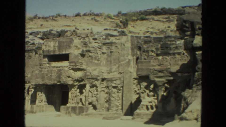 1974 India 8mm frame showing a carved rock-cut temple exterior with relief panels, a doorway, and sculpted stone figures.